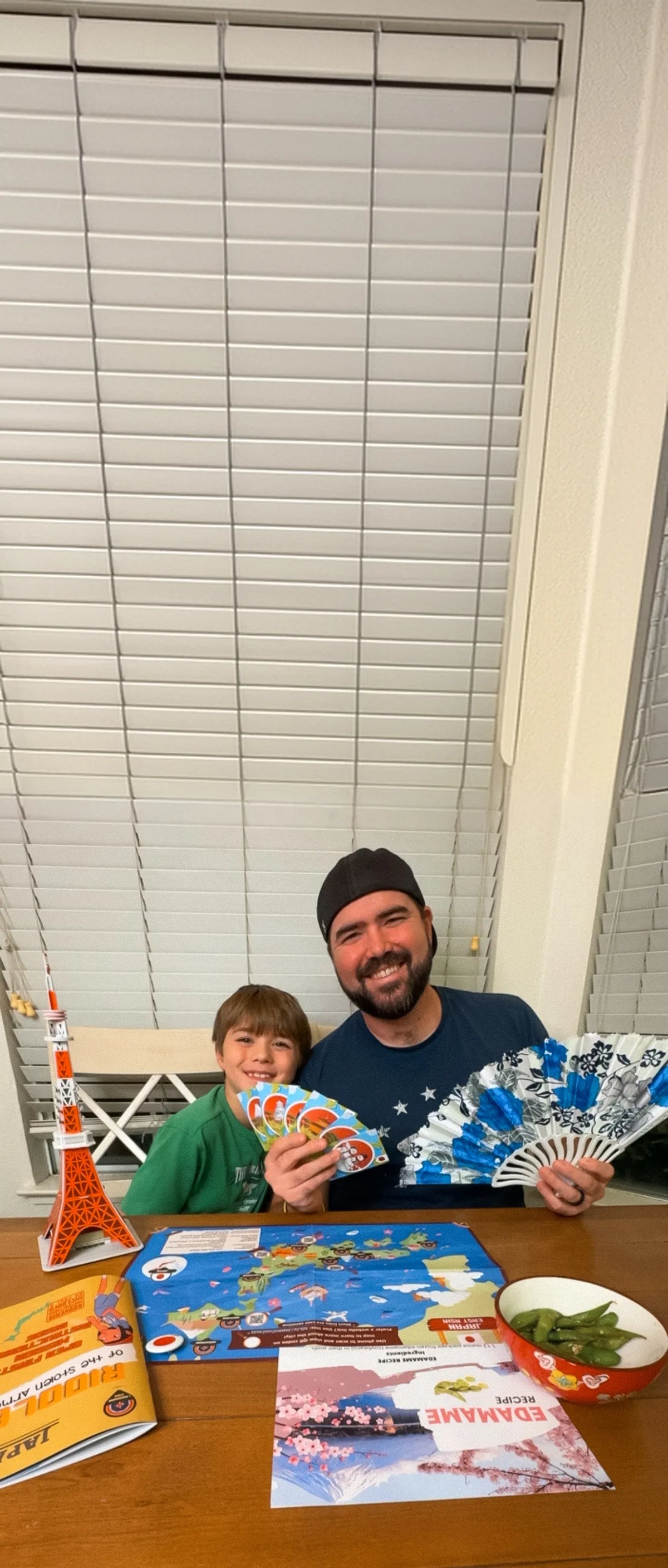 A father and son holding Japan mission cards and a paper fan next to a Tokyo Tower model and the mission map